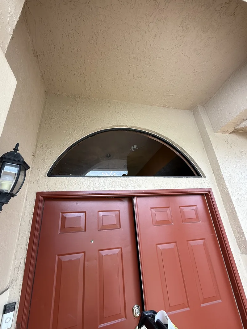 Curved transom opening beside a red front door with the old glass removed, showing the bare frame and stucco arch